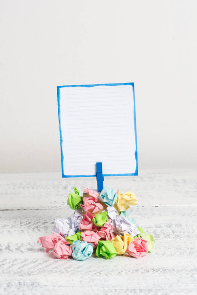 Square shaped reminder above a pile of crumpled papers fixed by a blue clothespin. Different sheets stacked under an empty colorful note. White wooden floor and white background. - Photo, Image