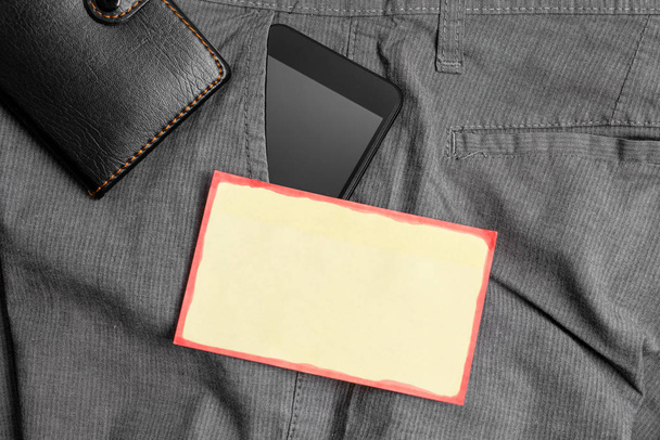 Smartphone device in the trousers front pocket with a wallet and yellow note paper. Modern gadget is placed inside man pants near notation sheet plus purse - Фото, изображение