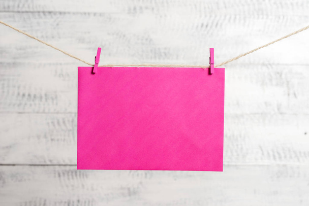 Rectangle shaped note colored paper clipped in a clothesline by clothespin in a white wooden background. Square empty reminder and office equipments supplies in a light wood desk. - Photo, image