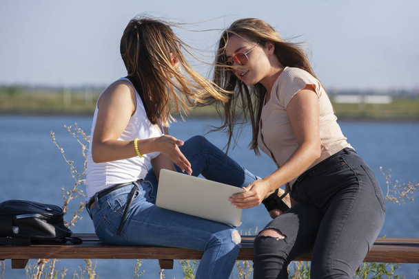 Two beautiful young caucasian girls looking amazed to a laptop while sitting outside in the park on a bench. - Photo, image