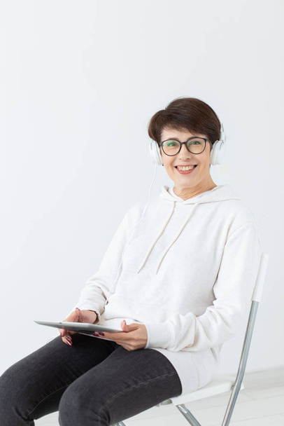 charmante schlanke Frau mittleren Alters mit Brille und Pullover sitzt auf einem Stuhl und hört ihr Lieblings-Online-Radio mit Hilfe eines Headsets und drahtlosem Internet und hält Tablet auf einem weißen - Foto, Bild