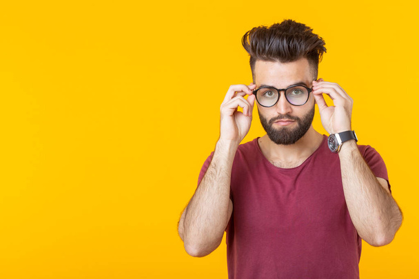 Young handsome guy is looking at his glasses while standing against a yellow background. The concept of poor vision and eye treatment. - Photo, Image