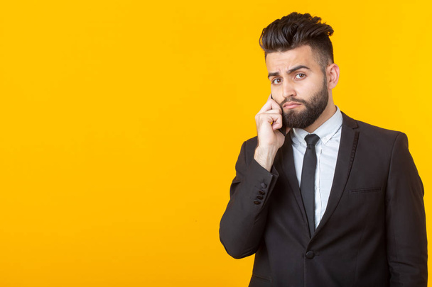 Cute young man with a beard in formal clothes talking on the phone posing on a yellow background with copy space. Concept of an informal meeting and business deal. - Фото, изображение