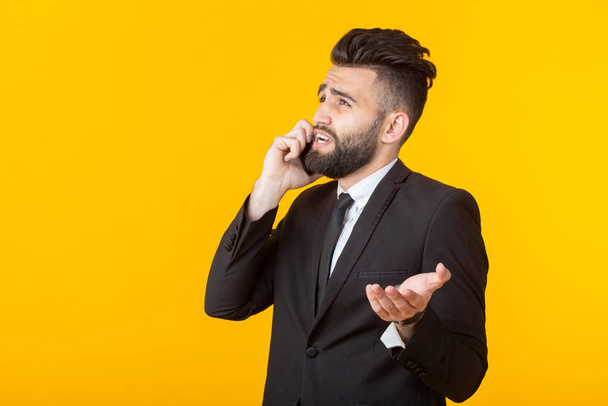 Милый молодой человек с бородой в формальной одежде разговаривает по телефону, позируя на жёлтом фоне. Концепция неформальной встречи и деловой сделки
. - Фото, изображение