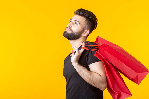 Side view of an upset young bearded stylish hipster man holding shopping bags posing on a yellow background. Concept of superfluous purchases. - Photo, Image