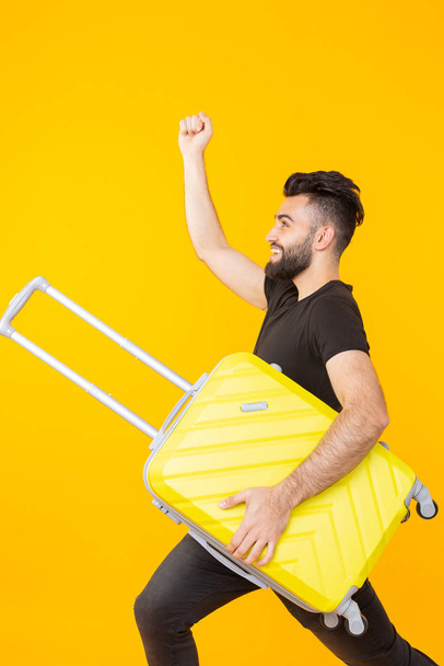 Cute young bearded hipster man holding a heavy yellow suitcase on a yellow background. Concept of heavy load for vacation travel. - Valokuva, kuva
