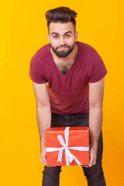 Портрет красивого молодого хипстера с бородой, позирующего на жёлтом фоне и держащего красную подарочную коробку. Концепция подарков и скидок
. - Фото, изображение