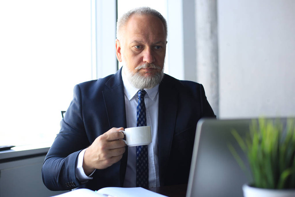 Mature businessman working on computer and drinking coffee in modern office. - Фото, зображення