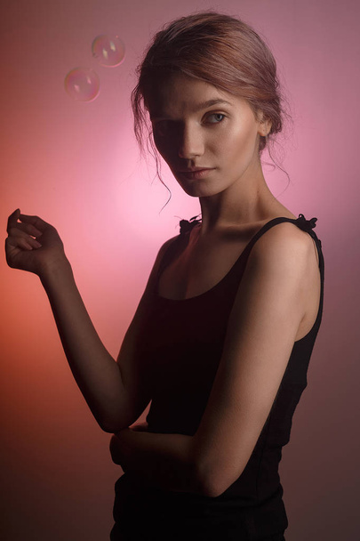 sensuele Kaukasische meisje in zwarte jurk, schaduw op haar gezicht op roze achtergrond. Twee zeepbellen vliegen rond haar hoofd. - Foto, afbeelding