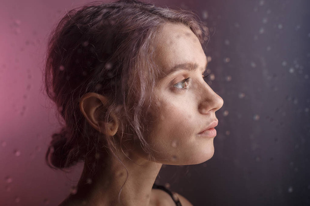 beautiful young girl looks away on purple background. blurry drops of water run down the glass in front of her face - Foto, Imagen