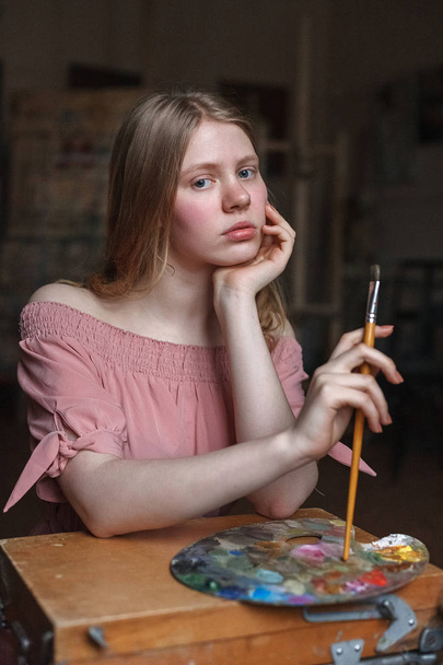 jeune fille blonde en robe rose repose soigneusement sa tête sur son bras et tient brosse à côté de la palette dans l'atelier d'art
 - Photo, image
