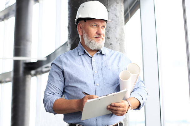 Confident mature business man in hardhat holding blueprint and looking away while standing indoors. - Valokuva, kuva