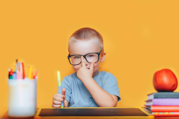 Feliz chico rubio inteligente está sentado en un escritorio con gafas y sonriendo. Listo para la escuela. De vuelta a la escuela. Manzana, bolígrafos y libros en el escritorio
 - Foto, Imagen