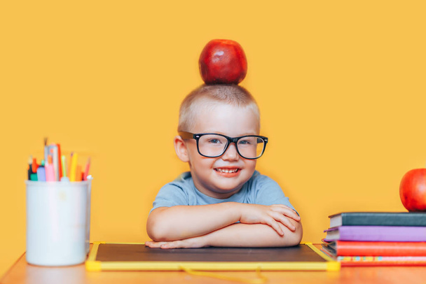 fröhlicher netter schlauer Junge sitzt am Schreibtisch und lächelt. Kind steht auf gelbem Hintergrund. Das erste Mal in die Schule. zurück zur Schule. Schüler in Gläsern mit Apfel auf dem Kopf - Foto, Bild