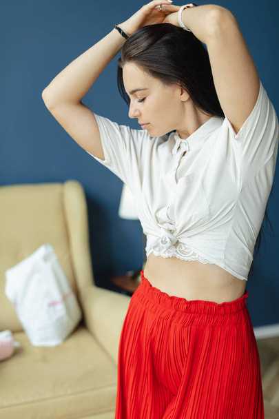 gorgeous brunette woman in red fashion pans with wide legs and white shirt sniffing armpits. fashion young woman indoor with black hair smelling herself. comfortable home concept. fine art woman - Foto, immagini