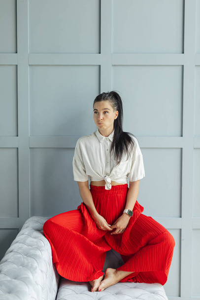 gorgeous brunette woman in red fashion pans with wide legs and white shirt. fashion young woman indoor with black hair on sofa without shoes. comfortable home concept. fine art woman portrait in - Φωτογραφία, εικόνα