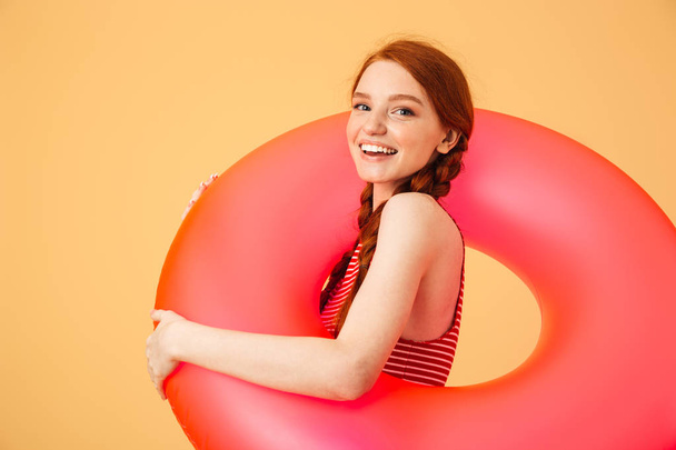 Smiling young beautiful redhead woman posing isolated over yellow background holding rubber ring. - Foto, Imagen