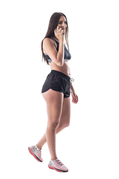 Πλευρική όψη του Happy χαμογελαστή ελκυστική γυναίκα γυμναστήριο σε αθλητικά είδη περπάτημα και μιλώντας στο τηλέφωνο. Ολόκληρο το σώμα απομονώνεται σε λευκό φόντο.  - Φωτογραφία, εικόνα