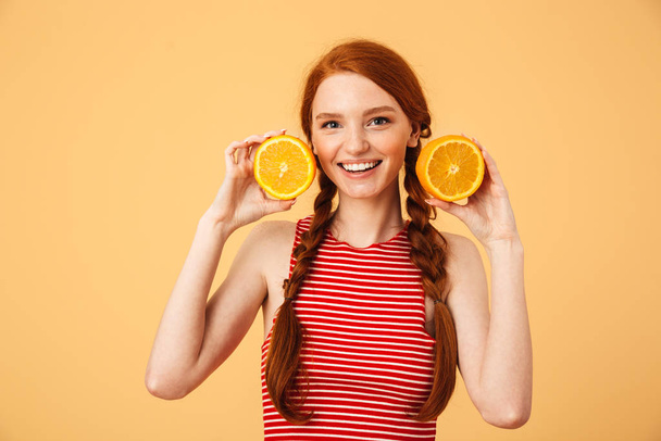 Веселая молодая красивая рыжая женщина позирует изолированно на желтом фоне держа оранжевый
. - Фото, изображение