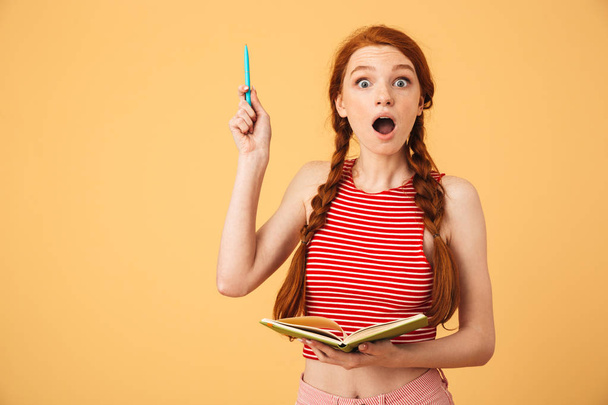 Shocked  young beautiful redhead woman posing isolated over yellow background holding book reading have an idea. - Foto, Imagen