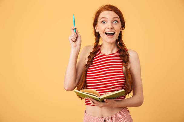 Surprised  young beautiful redhead woman posing isolated over yellow background holding book reading have an idea. - Fotografie, Obrázek