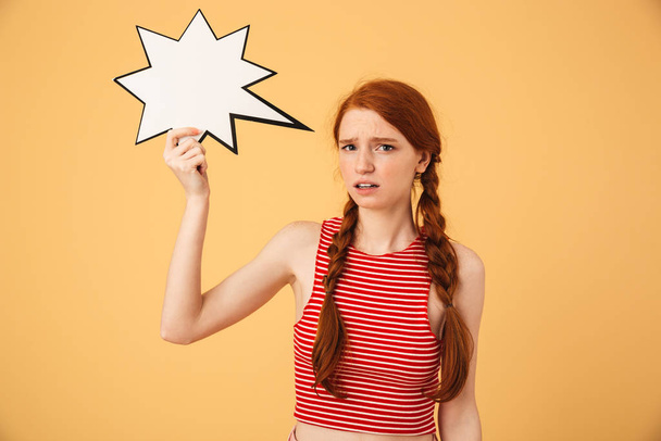 Confused young beautiful redhead woman posing isolated over yellow background holding speech bubble. - Φωτογραφία, εικόνα