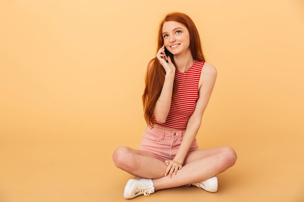 Cheerful young beautiful redhead woman posing isolated over yellow background talking by mobile phone. - Фото, изображение