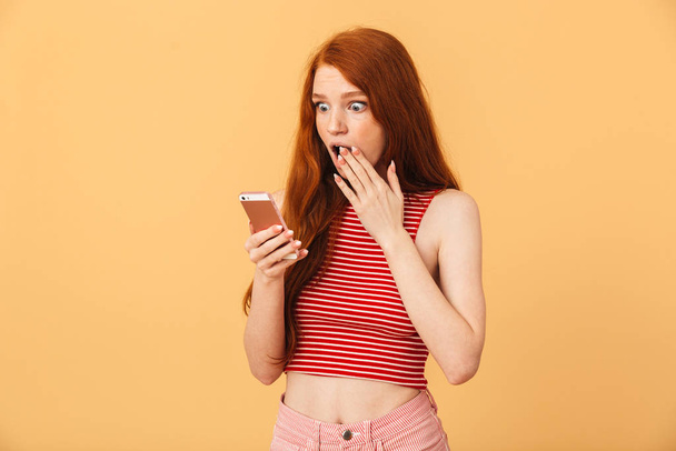 Shocked emotional cute young pretty redhead woman posing isolated over yellow background using mobile phone chatting. - Photo, Image