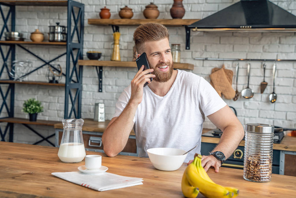 Ωραίος όμορφος μουσάτος άνθρωπος κάθεται στην κουζίνα με δημητριακά πρωινό και γάλα, καφέ και μπανάνες. Διαβάζω τον πρωινό τύπο, μιλάω στο κινητό και χαμογελάω. Υγιεινά φαγητά. Διατροφή. - Φωτογραφία, εικόνα