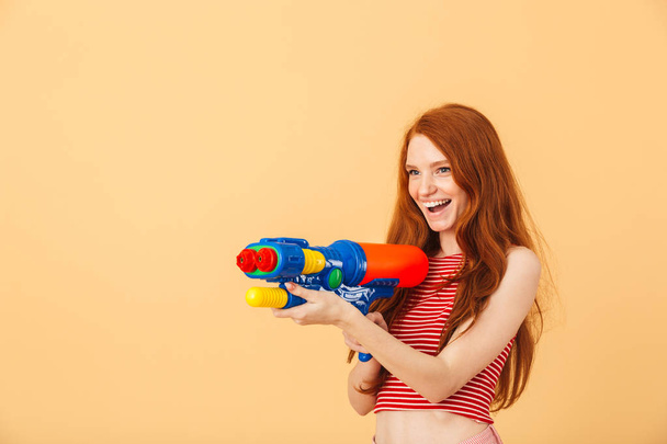 Excited young beautiful redhead woman posing isolated over yellow background holding water gun toy. - Foto, immagini