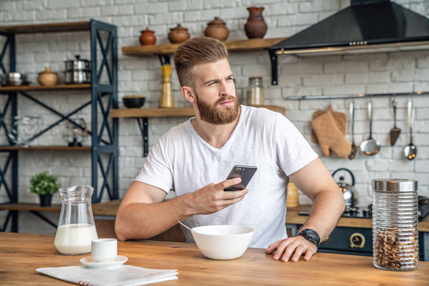 Hombre barbudo guapo bien parecido está sentado en la cocina tomando cereales de desayuno y leche, café y plátanos. sosteniendo su teléfono celular y mirando a un lado. Comida saludable. Dieta. Estilo de vida saludable
. - Foto, Imagen