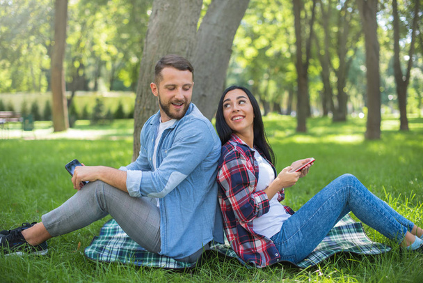 loving couple sitting on a plaid blanket in the park and use the phone, handsome guy and girl - Photo, image