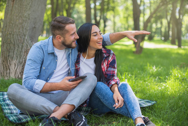 Liebendes Paar sitzt auf einer karierten Decke im Park und benutzt das Telefon, gutaussehender Typ und Mädchen - Foto, Bild