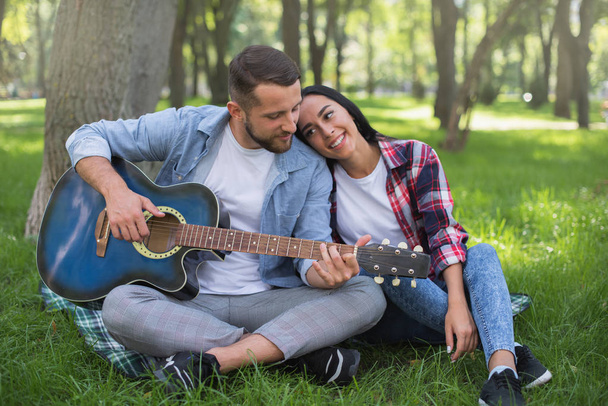 Mann und Mädchen spielen Gitarre im Park, sitzen auf dem Gras neben dem Baum - Foto, Bild