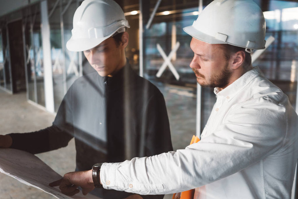 Safety inspector and build worker inside building under construction. Engineer hold blueprint in hand and discussing project with builder on construction site - Фото, изображение