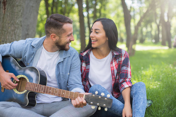 guy and girl play the guitar in the park, sit on the grass near the tree - Photo, Image