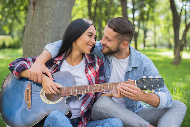 cara e menina jogar a guitarra no parque, sente-se na grama perto da árvore
 - Foto, Imagem