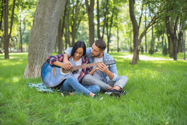 τύπος και κορίτσι παίζουν κιθάρα στο πάρκο, κάθονται στο γρασίδι κοντά στο δέντρο - Φωτογραφία, εικόνα