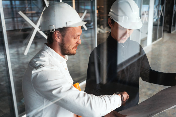 Two building engineers wearing safety hard hat discussing blueprint on construction site. Worker and inspector have meeting inside building under construction - Фото, изображение