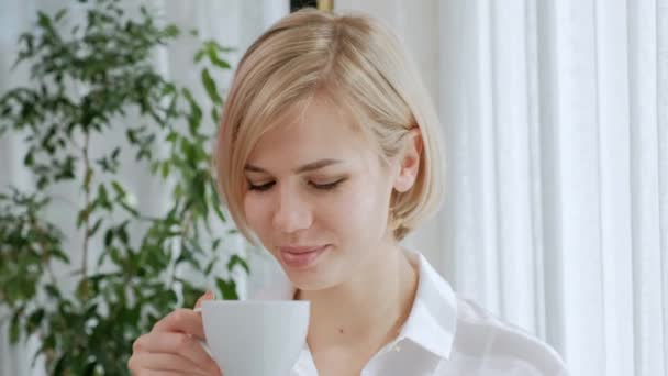 Egy fiatal, gyönyörű szőke nő, rövid hajú, szemüveg italok kávét egy fehér pohár egy könnyű lakásban. - Felvétel, videó