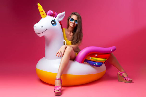 Portrait pf a young beauty brunette woman posing in fashion yellow  swimsuit on a white inflatable unicorn on pink isolated backgroung . Stylish sensual girl with an ideal body in bikini.  - Zdjęcie, obraz