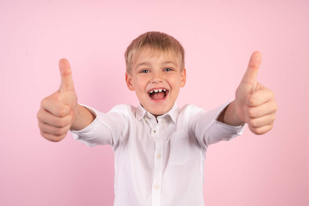Portret van kleine schattige jongen met plezier en glimlachend. Duim omhoog. studio portret over roze achtergrond. dragen jeans en wit overhemd. - Foto, afbeelding