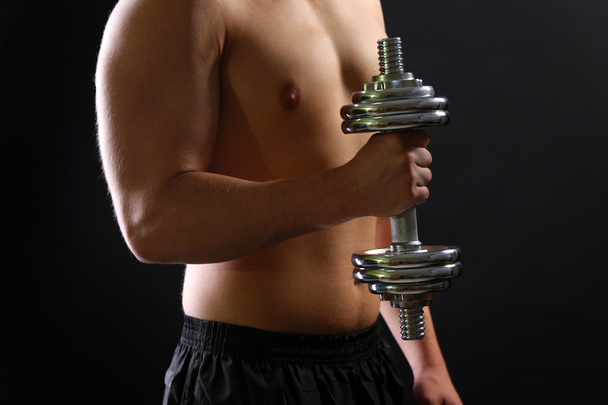 Handsome young muscular sportsman execute exercise with dumbbell on dark background - Zdjęcie, obraz