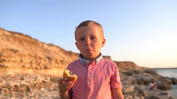 Zbliżenie, chłopiec dziecko jedzenia kok na brzegu. Prawdziwe pozytywne emocje, nagrania wideo w czasie rzeczywistym, chłopiec na tle wybrzeża morskiego, 4K - Materiał filmowy, wideo