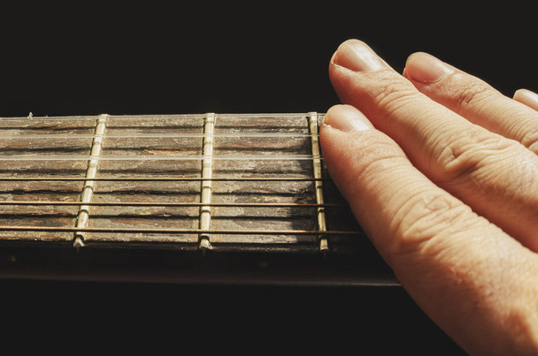 Пальцы приглушают звук струн старой акустической гитары, изолированной на черном фоне. Шея старой акустической гитары с ладами и ржавыми нейлоновыми струнами, изношенная и пыльная
.  - Фото, изображение