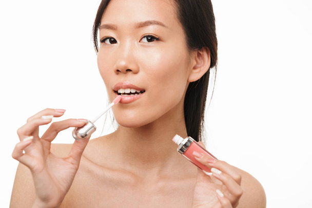 Beautiful asian woman posing isolated over white wall background holding makeup lipstick lip gloss. - Фото, изображение