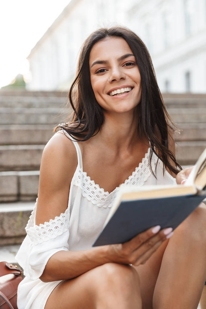 Portrait of smiling young woman reading book while sitting on st - Fotoğraf, Görsel