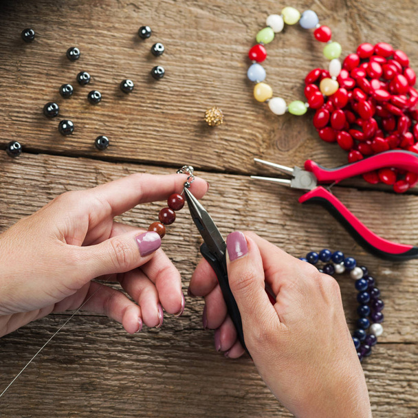 Sieraden maken. Het maken van armband van kleurrijke kralen. Vrouwelijke handen met een gereedschap op een ruwe houten tafel. Selectieve focus.  - Foto, afbeelding