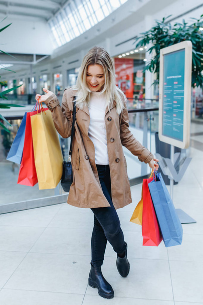 Ganzkörperporträt einer fröhlichen jungen blonden Frau in braunem Mantel nach dem Einkaufen. Schöne Frau mit bunten Einkaufstaschen, die in einem Einkaufszentrum steht. Shopper, Verkauf, Einkaufszentrum - Foto, Bild