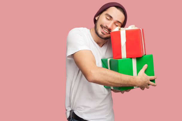 happy satisfied handsome bearded young hipster man in white t-shirt and casual hat hugging two present boxes on pink background - Foto, afbeelding